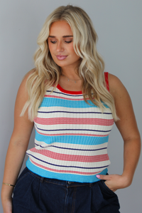 A woman modeling a sleeveless ribbed knit top with horizontal stripes in red, light blue, navy, and white, paired with dark wide-leg jeans and metallic rose gold flats, standing indoors against a white wall.
