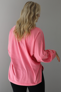 Woman wearing a pink oversized long sleeved top that has a high neckline, exposed seam details, split hem details on either side, and is lightweight. The top is modeled with a pair of fitted black leggings. The photo is modeled indoors against a white wall. The photo shows the back of the top. 