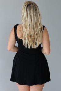 Woman wearing a black mini dress with thick straps, a fitted bodice, and flowy skirt indoors against a white wall. The photo shows the back of the dress. 