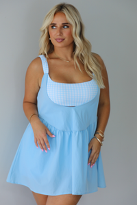 A woman modeling a light blue sleeveless mini dress with a white gingham bodice and a flared skirt, paired with brown slide sandals, standing indoors against a white wall.
