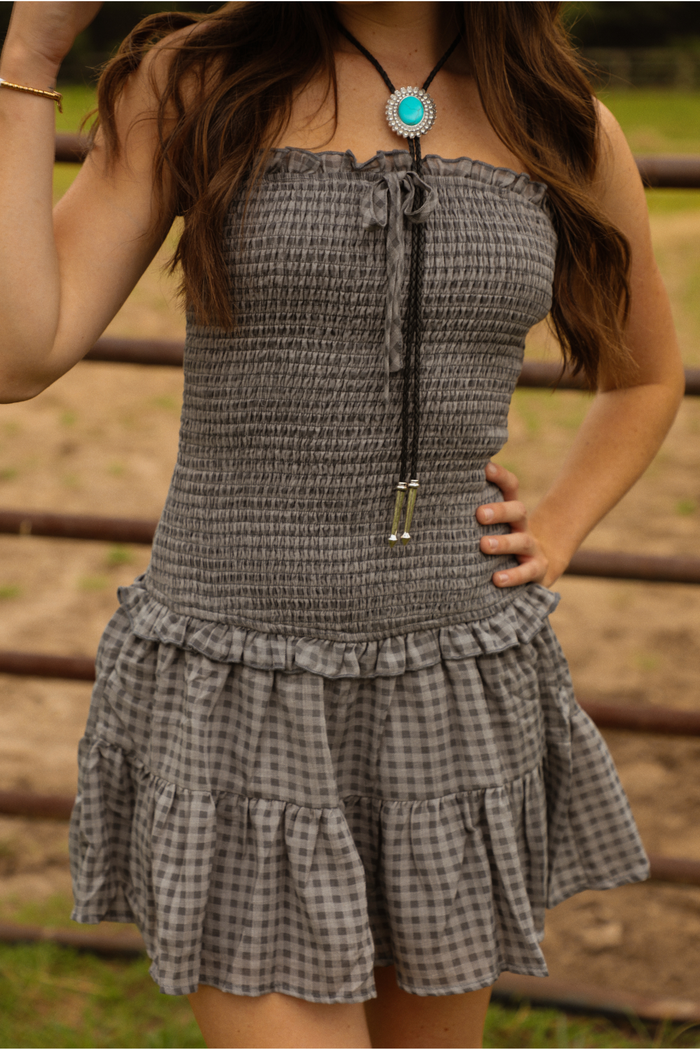Woman wearing a charcoal and grey gingham patterned strapless dress in a pasture, with a metal gate in the background. The dress is strapless, has a smocked drop-waist design, and a ruffled skirt. The dress is paired with black, silver, and turquoise bolo tie. 