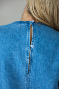 Woman wearing a v-neck, short sleeved, denim mini dress with functional pockets on either side. The photo is taken indoors against a white wall and shows the back of the dress with the button closures at the nape of the neck.