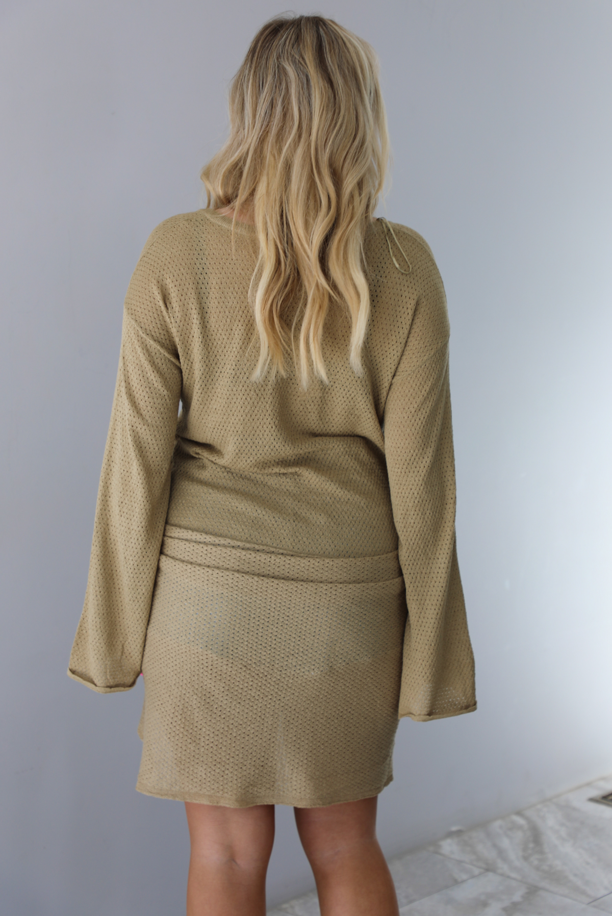 Woman wearing a camel long-sleeve dress that has a tie detail in the front.  The photo is taken indoors against a white wall. The photo shows the back of the dress. 