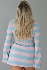 Woman wearing a pink and blue striped dress that has long sleeves and a mini length. The photo is taken indoors against a white wall and shows the back of the dress. 