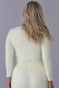 Woman wearing a light yellow three piece set featuring a long sleeved top, long, flared hem pants, and a long, matching cardigan. The photo is taken indoors against a white wall and shows a closeup of the back of the top. 