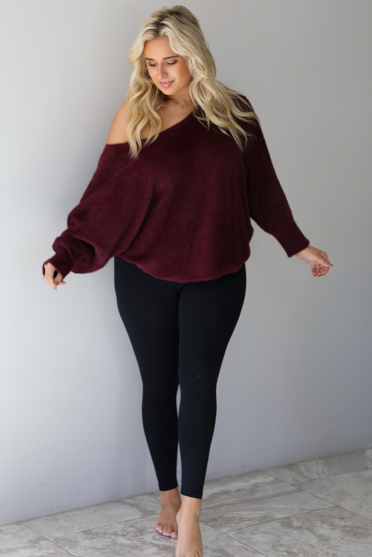 A woman wearing a maroon oversized long sleeved sweater, worn off the shoulder, paired with black leggings, indoors against a white wall with white marble flooring.