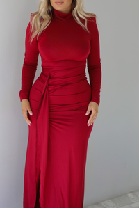 Woman wearing a red maxi dress that has a high neckline, shoulder pads, a fitted bodice, long sleeves, with a split hem detail on the left side. The photo is taken indoors against a white wall with white marble flooring. The photo shows a closeup detail to the waist of the dress.