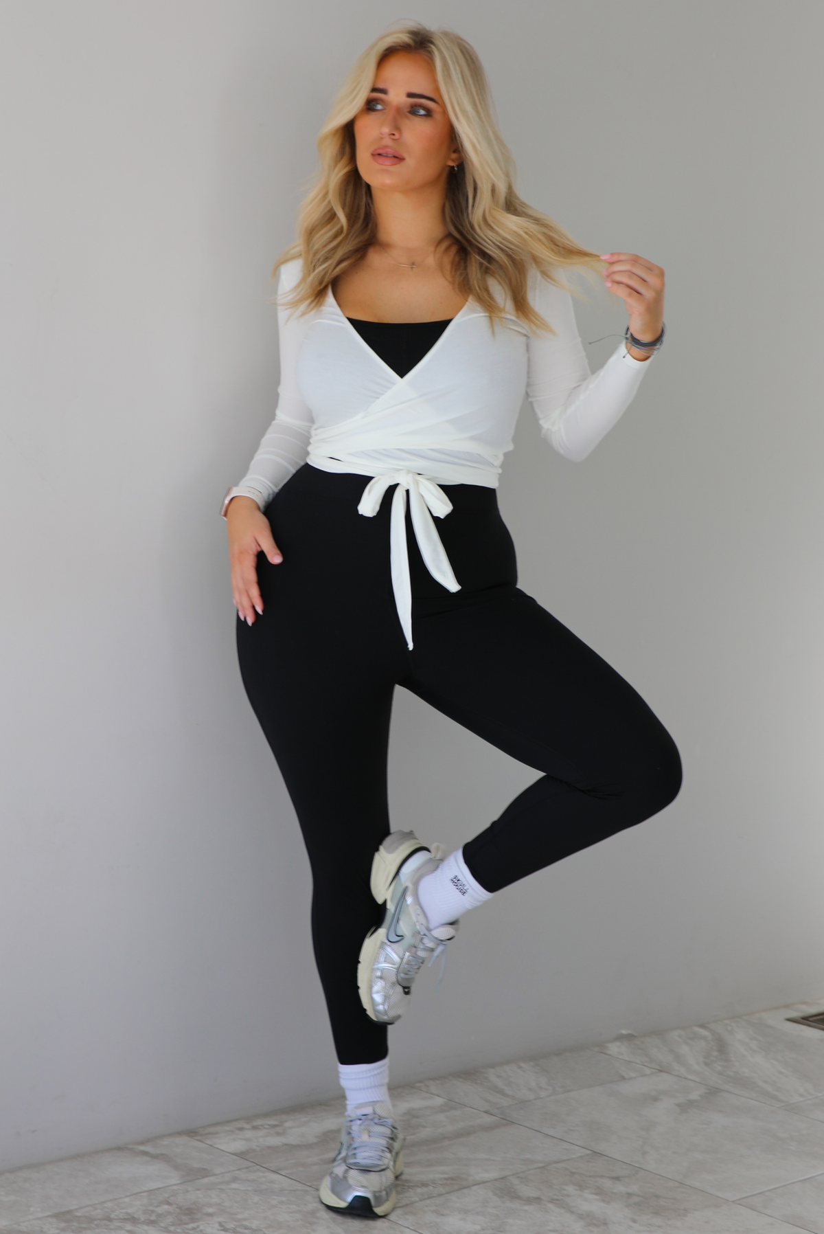 Woman wearing a white cropped wrap style fitted top over a black fitted jumpsuit. The outfit is paired with white ankle socks and silver sneakers. The photo is taken indoors against a white wall with white marble flooring. 