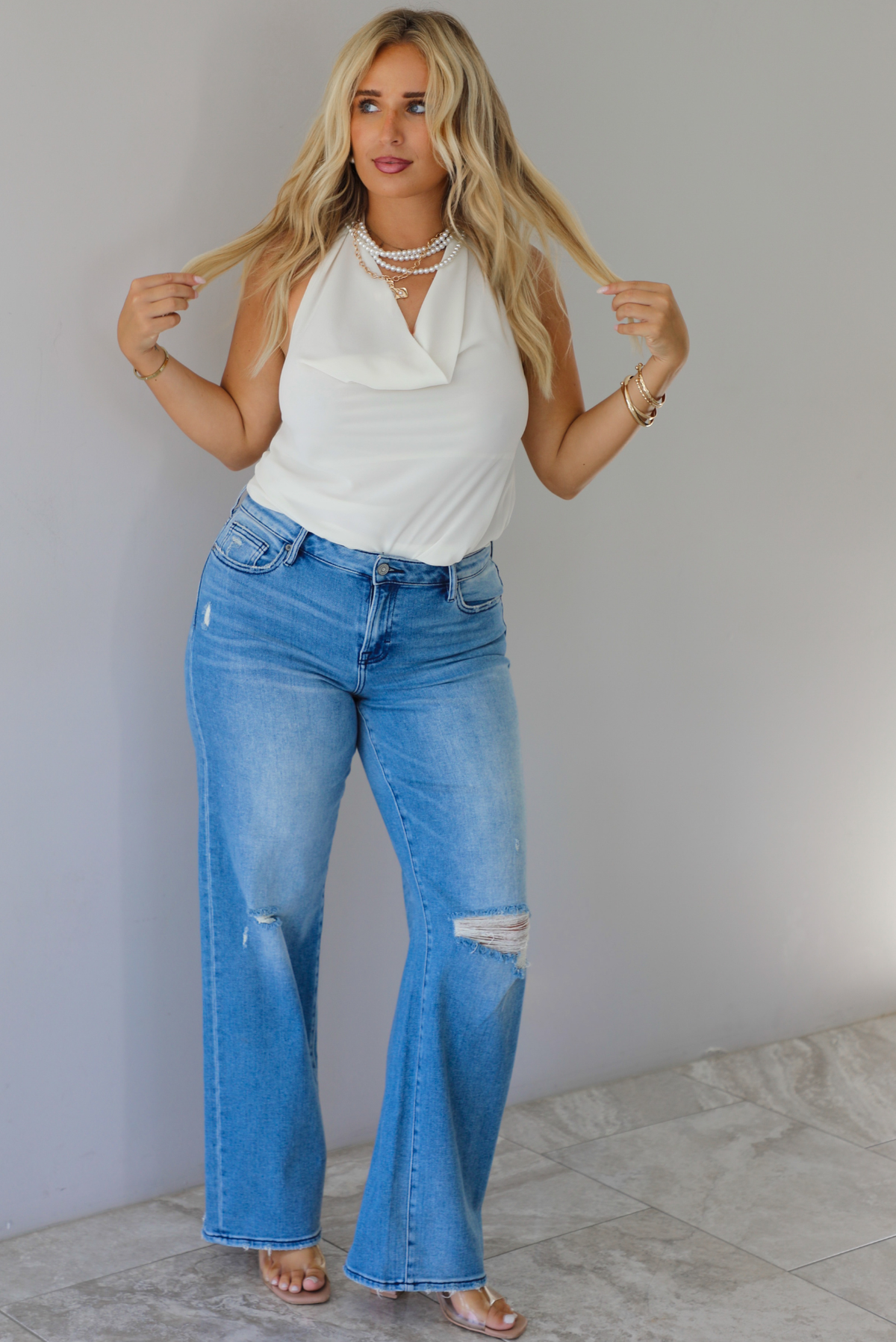 A woman modeling a cream sleeveless halter top and light-wash blue distressed flare jeans, standing indoors against a white wall with white marble flooring. 