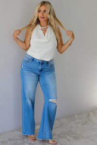 A woman modeling a cream sleeveless halter top and light-wash blue distressed flare jeans, standing indoors against a white wall with white marble flooring. 
