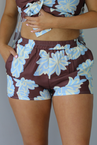 Woman wearing a brown, blue, and off white floral two piece set featuring a vest style top with functional button closures in the front and shorts. The photo is taken in front of a white wall and shows a closeup of the shorts, which have an elastic waistline and functional pockets on either side. 