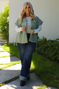Woman model wearing a sage colored oversized vest paired with a white lace long sleeved top and dark denim outdoors.  The vest has functional pockets on either side, functional button closures in the front, and an oversized fit. 