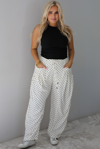 A woman wearing a black high neck tank paired with sand white pants with a black polka dot print pattern throughout, standing indoors against a white wall with white marble flooring. 