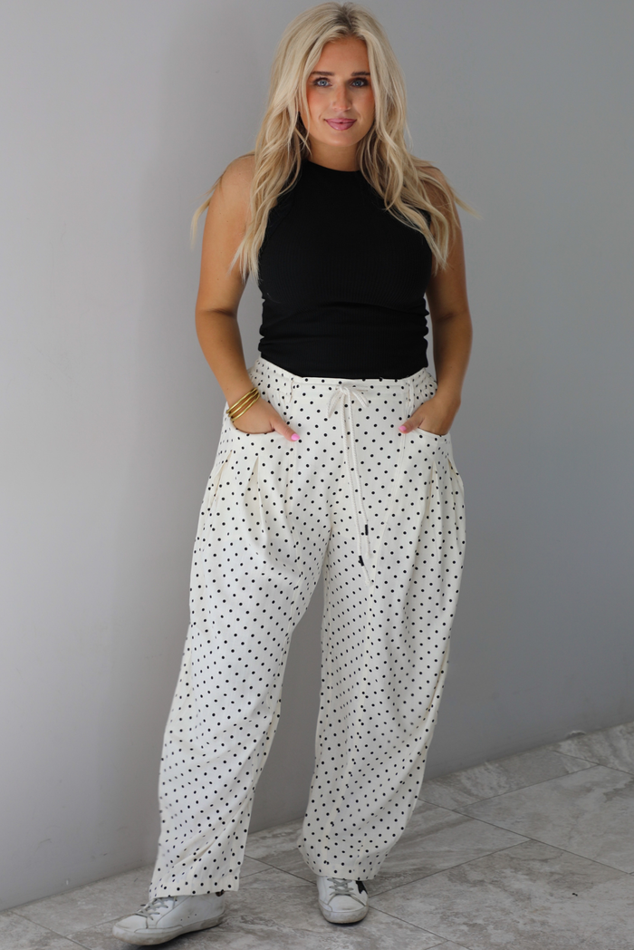 A woman wearing a black high neck tank paired with sand white pants with a black polka dot print pattern throughout, standing indoors against a white wall with white marble flooring. 
