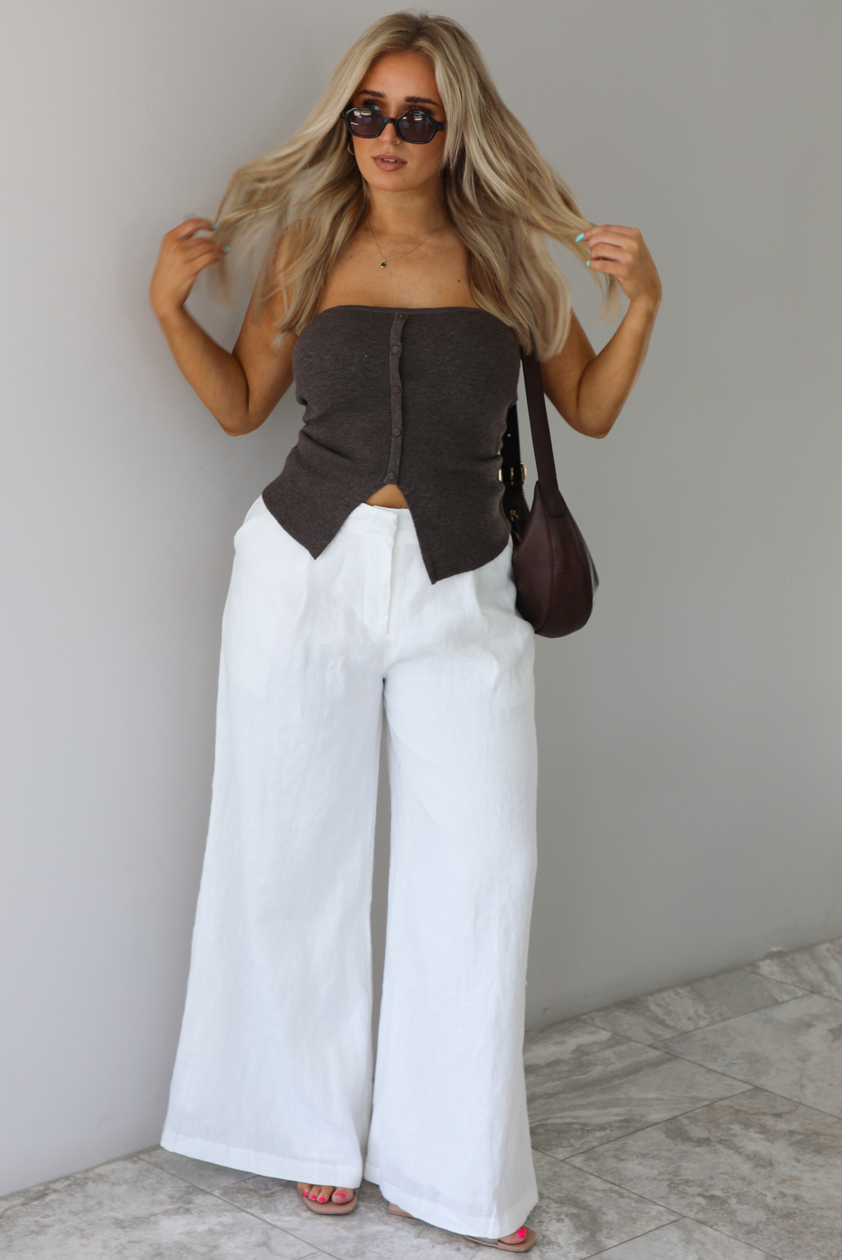 Woman wearing a strapless brown top with non-functional button closures in the front with  a split hem detail, and high waisted, flared hem white linen pants with a brown shoulder bag. The photo is taken indoors against a white wall with white marble flooring. 