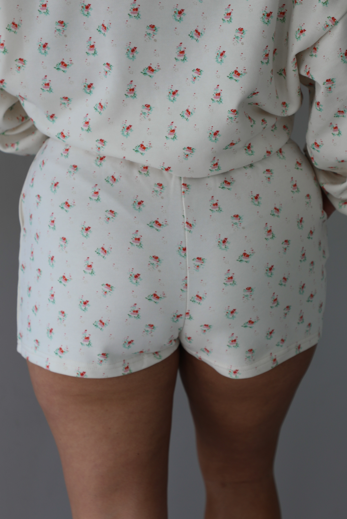 Woman wearing a two piece cream set that has a floral design throughout featuring a sweatshirt and sweat-shorts indoors against a white wall. The photo shows a closeup of the back of the shorts. 