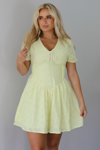 Woman wearing a light yellow mini dress with a floral eyelet design throughout, v-neckline, short sleeves, fitted drop waist bodice, and a flowy mini length skirt indoors against a white wall.