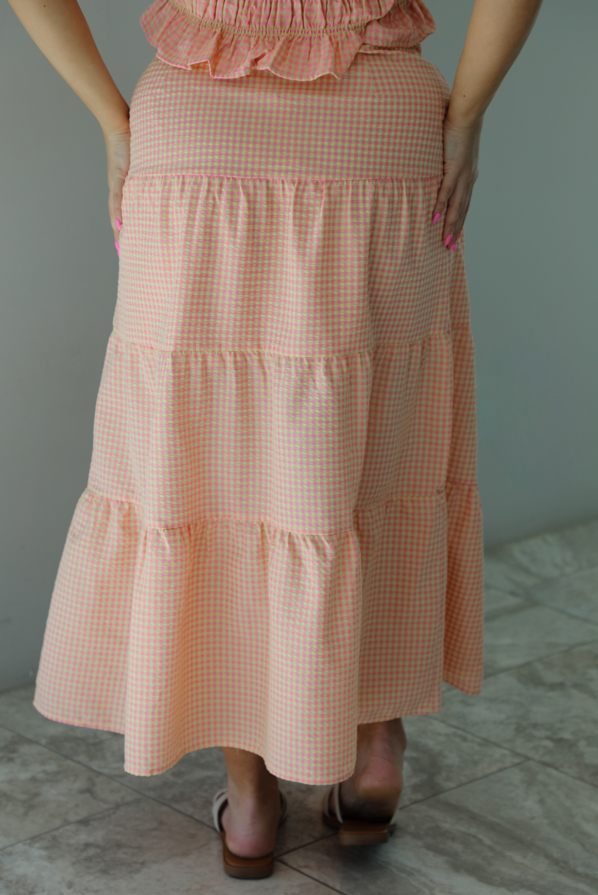 Woman wearing a pink and pale yellow gingham print two piece set featuring a smocked top and high waisted, maxi length skirt. The photo is taken indoors against a white wall and shows a closeup of the back of the skirt, which is high waisted and has a maxi length.