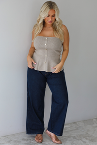 A woman wearing a beige strapless button-front peplum top and dark denim wide-leg jeans, standing indoors against a white wall with white marble flooring. 