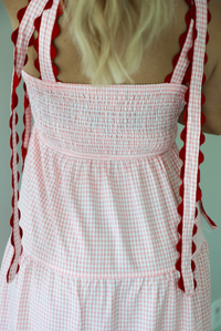 Woman wearing a pink and white gingham  maxi dress with thick straps, a fitted bodice, flowy skirt, and crocheted cherry details on the front indoors against a white wall. The photo shows the back of the dress, which has a smocked detail at the waistline. 