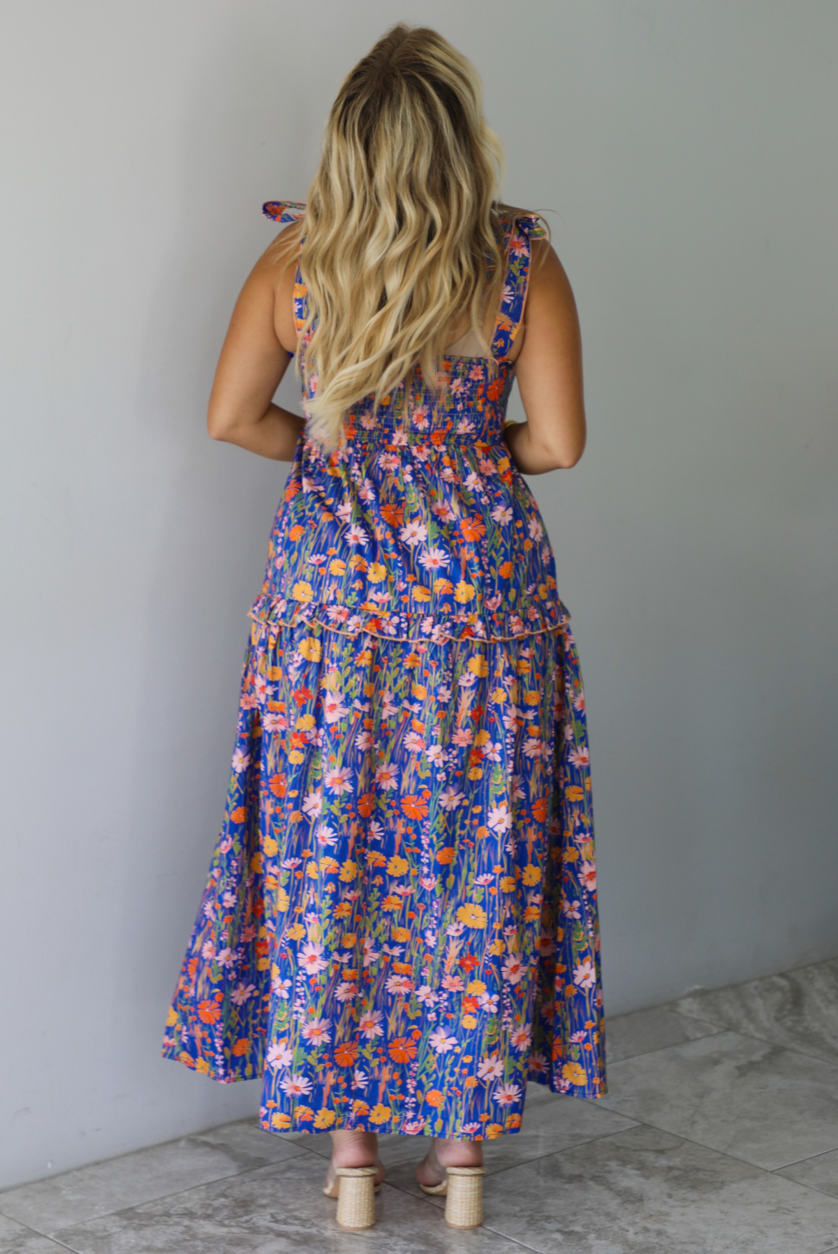 A woman shown from behind wearing a sleeveless tiered navy blue maxi dress with a colorful floral print with a fitted bodice and flowy skirt, standing indoors against a white wall with white marble flooring. 