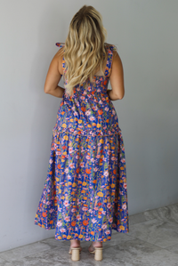 A woman shown from behind wearing a sleeveless tiered navy blue maxi dress with a colorful floral print with a fitted bodice and flowy skirt, standing indoors against a white wall with white marble flooring. 