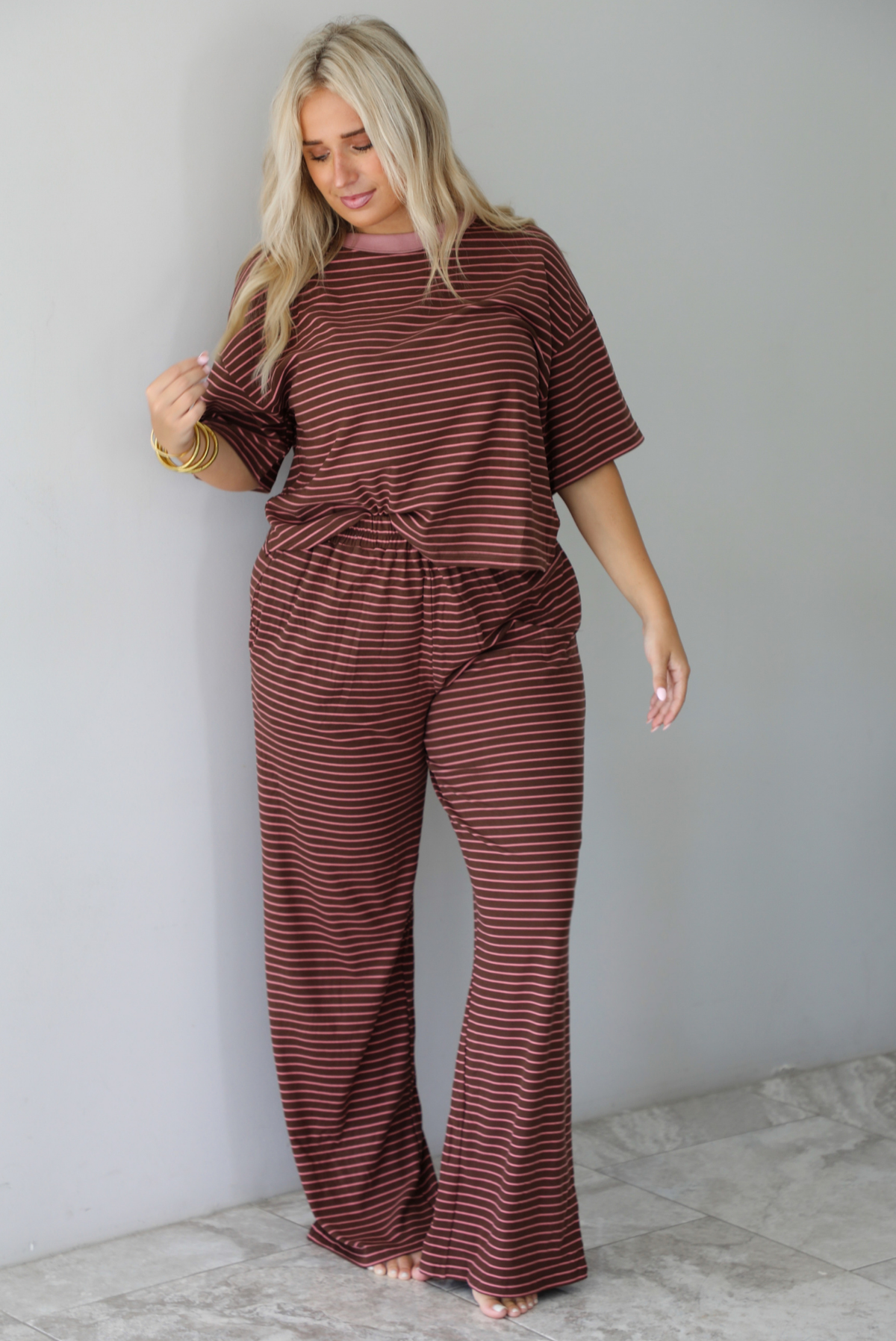 A woman wearing a brown and pink striped two piece set featuring a slightly cropped tee and high waisted pants, with the top slightly tucked into the pants, standing indoors against a white wall with white marble flooring. 