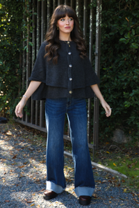 Woman model outdoors wearing a charcoal grey colored cape cardigan with functional button closures in the front and dark wash denim. 