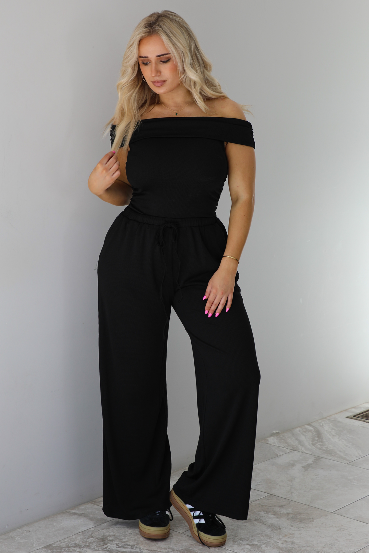 Woman wearing a two piece set featuring an off the shoulder top and high waisted pants. The photo is taken indoors against a white wall with white marble flooring. 