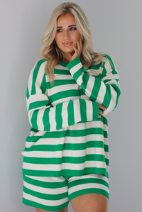 Woman wearing a green and white striped set featuring an oversized long sleeved top and high waisted shorts. The photo is taken indoors against a white wall.