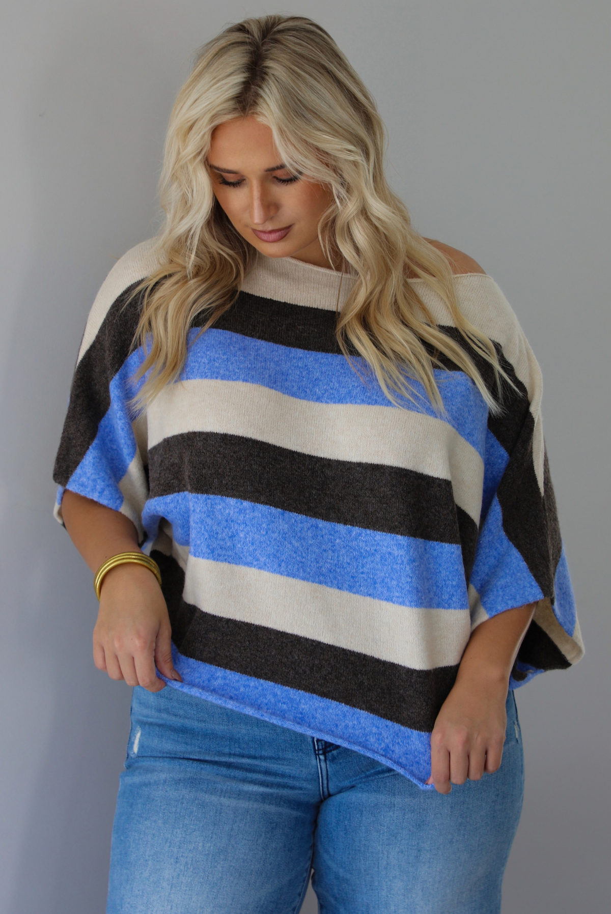 A woman wearing a cream, brown,, and blue striped knit top that has a boat-neck design, and quarter length flowy sleeves, paired with a pair of medium wash denim jeans, standing indoors against a white wall.