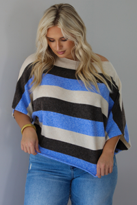 A woman wearing a cream, brown,, and blue striped knit top that has a boat-neck design, and quarter length flowy sleeves, paired with a pair of medium wash denim jeans, standing indoors against a white wall.