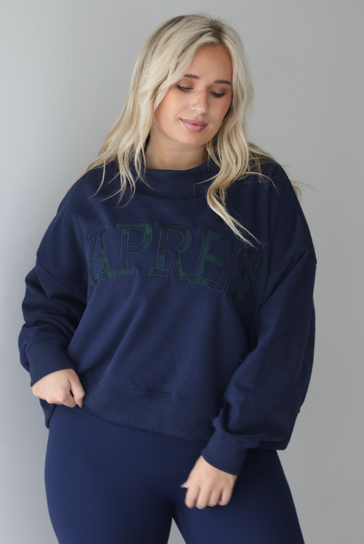 A woman wearing a navy sweater with Apreś written on the front in collegiate lettering in a plaid print, paired with a matching two piece navy set, standing indoors against a white wall.