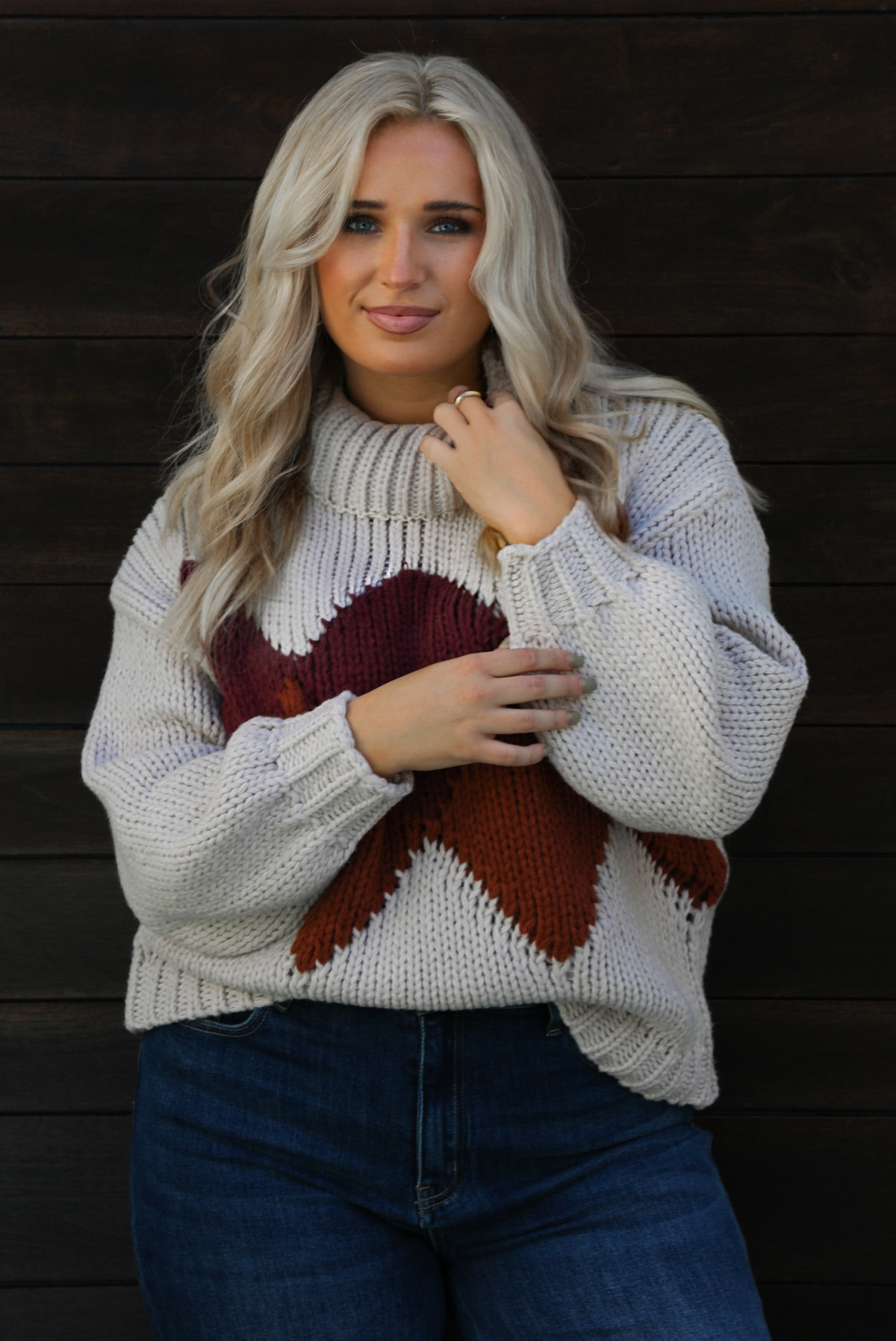 Close up of a woman model wearing a chunky knit turtle neck sweater with dark denim jeans outdoors in front of a dark wood door. 