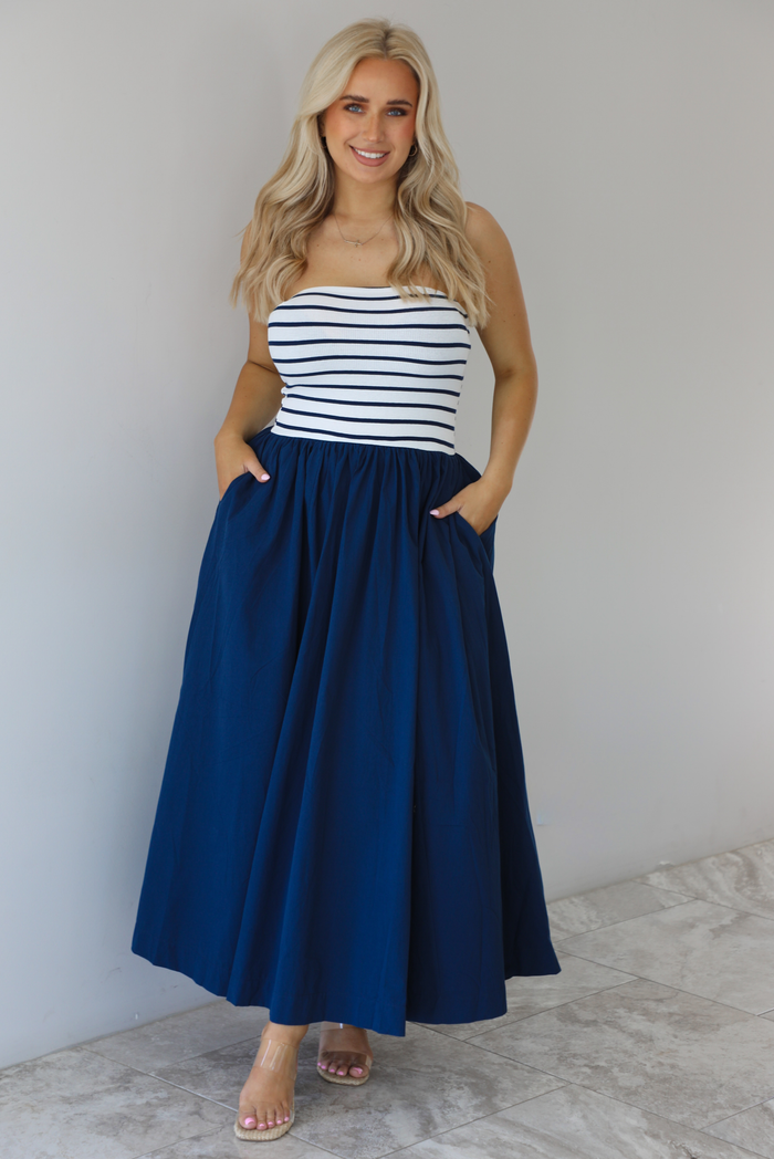 Woman wearing a maxi dress featuring a navy and white striped bodice and navy, maxi length skirt. The photo is taken indoors against a white wall with white marble flooring. 