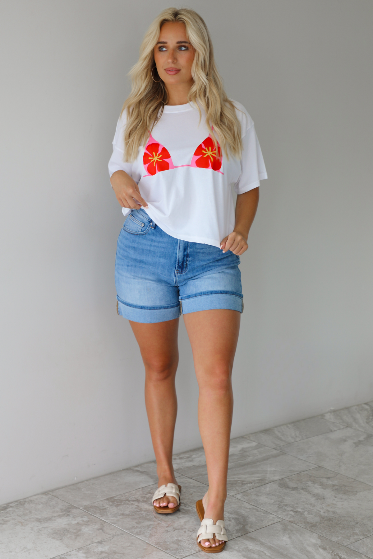 Woman wearing a white cropped boxy tee with a pink, red, and yellow bikini graphic on the front with a pair of medium wash denim shorts indoors against a white wall with white marble flooring.