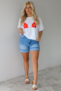 Woman wearing a white cropped boxy tee with a pink, red, and yellow bikini graphic on the front with a pair of medium wash denim shorts indoors against a white wall with white marble flooring.