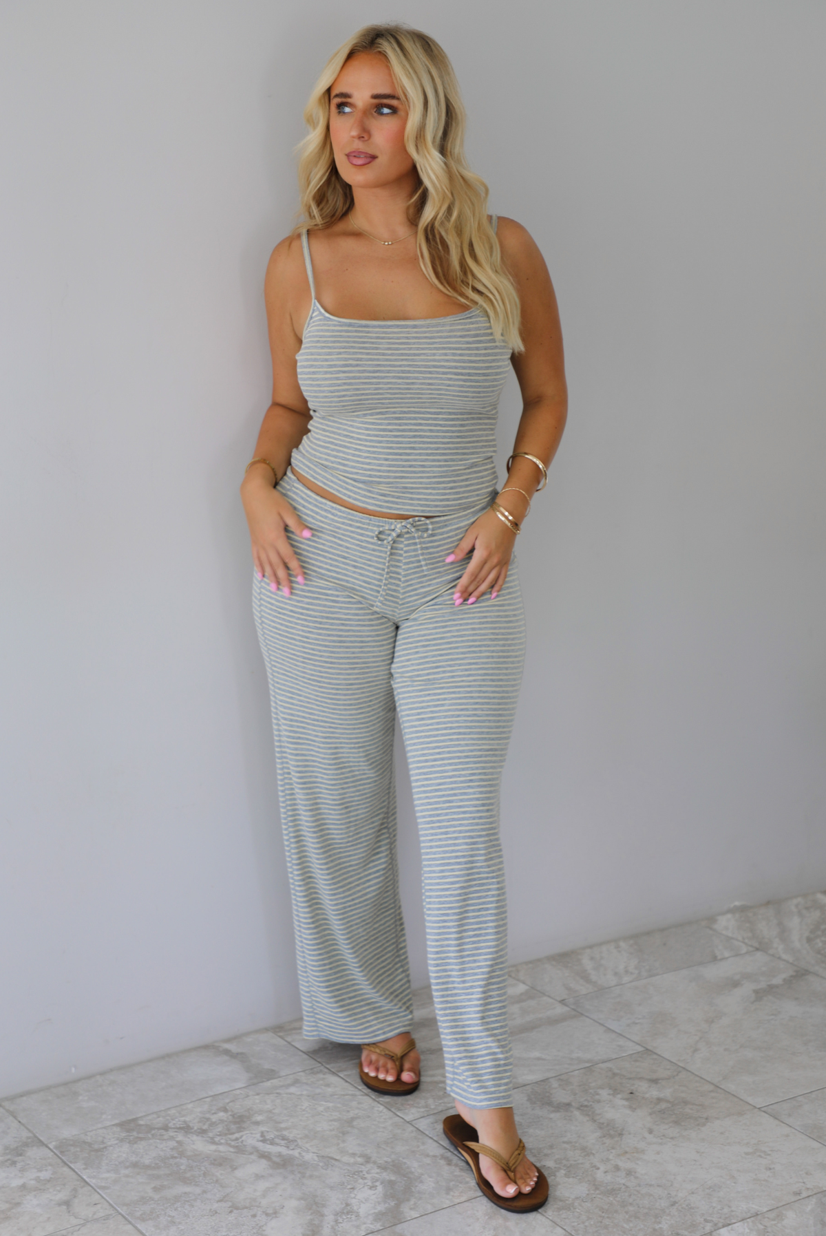 A woman modeling a matching two-piece loungewear set consisting of a tank top top and wide-leg drawstring pants, both in a light grey and yellow striped pattern, standing indoors against a white wall with white marble flooring. 