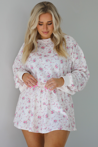 Woman wearing a pale pink two piece set that has a pink floral print pattern throughout. The set featuring an oversized long sleeved top and high waisted skirt. The photo is taken indoors against a white wall.