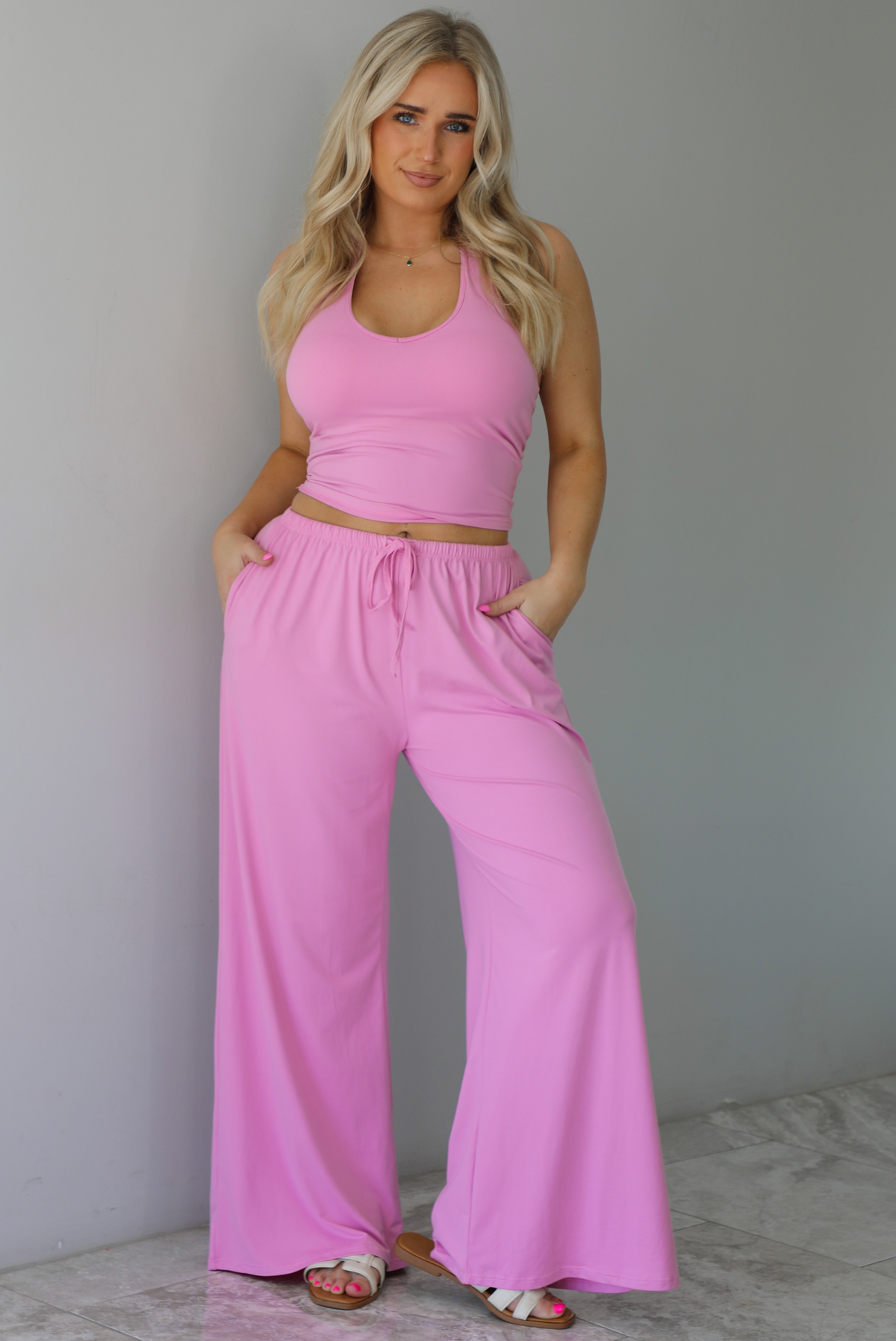 Woman wearing a two pieced pink set featuring a halter top and comfy pants with a flared hem detail. The photo is taken indoors against a white wall with white marble flooring. 
