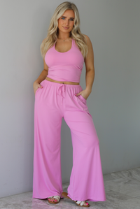 Woman wearing a two pieced pink set featuring a halter top and comfy pants with a flared hem detail. The photo is taken indoors against a white wall with white marble flooring. 