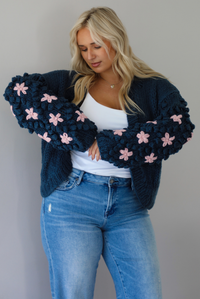 A woman wearing a navy oversized cardigan with a pink embroidered floral design on the sleeves, paired with a white tank, mid-rise medium wash denim jeans, standing indoors against a white wall. The model is holding both arms up to display the embroidered floral details on the sleeves. 