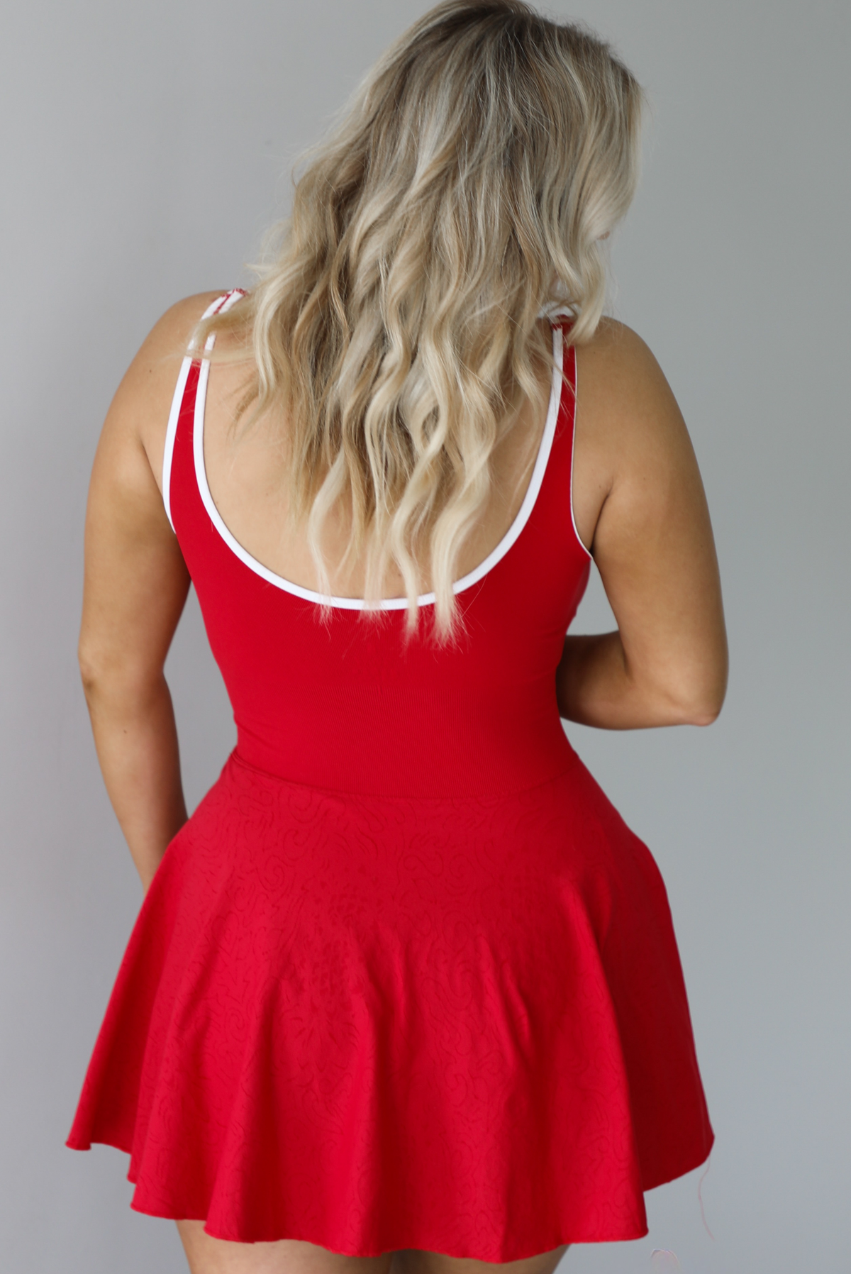 Woman wearing a red athleisure dress that has white trim, a fitted bodice, and flowy skirt. The photo shows a closeup of the back of the dress. The photo is taken indoors against a white wall.