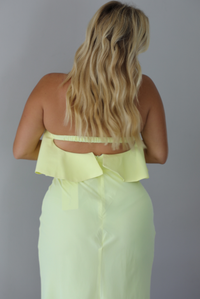 A woman viewed from behind wearing a pale yellow ruffled strapless maxi dress, standing indoors against a white wall. The photo shows a closeup of the upper-back of the dress, showing the zipper closure. 