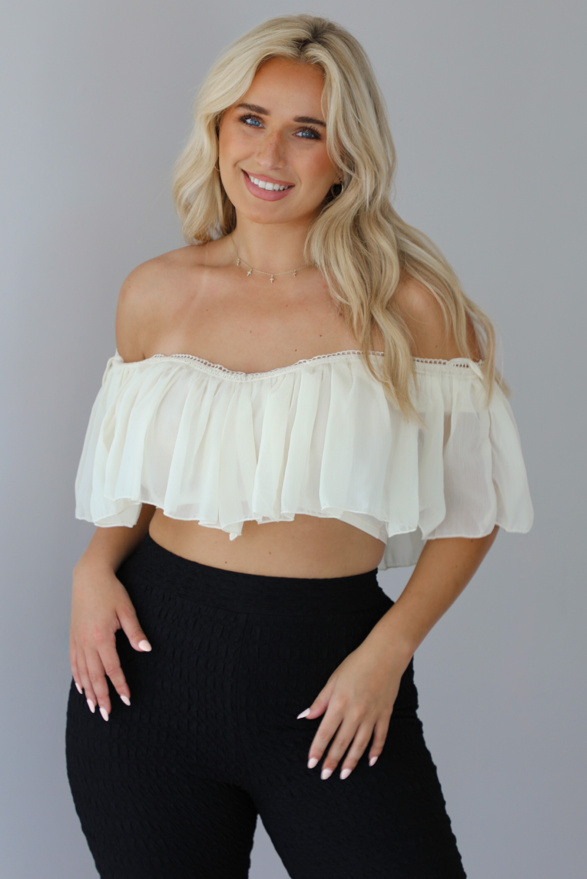 Woman wearing a cream ruffled cropped top with high waisted black, textured capri pants indoors against a white wall. The photo shows a closeup of the top, which is being worn off the shoulder. 