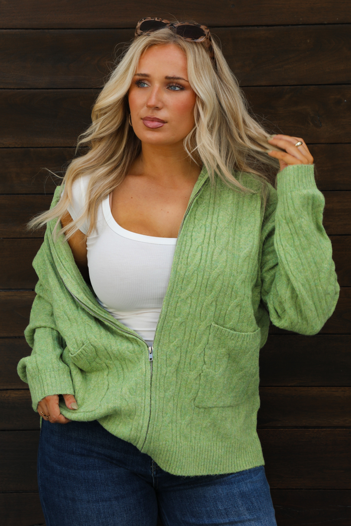 Woman model wearing a green cable knit zip-up cardigan with dark denim jeans outdoors against a dark brown wooden door. The cardigan is zipped and she's wearing a white top underneath. The model is wearing a large.