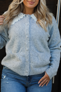 Close up of a woman model wearing a heather grey cardigan with a white lace bib at the neckline, has functional button closures in the front, functional pockets in the front, and a relaxed fit. The cardigan is paired with a medium wash denim and tan ugg boots. The photo is taken outdoors against a dark brown wooden door.