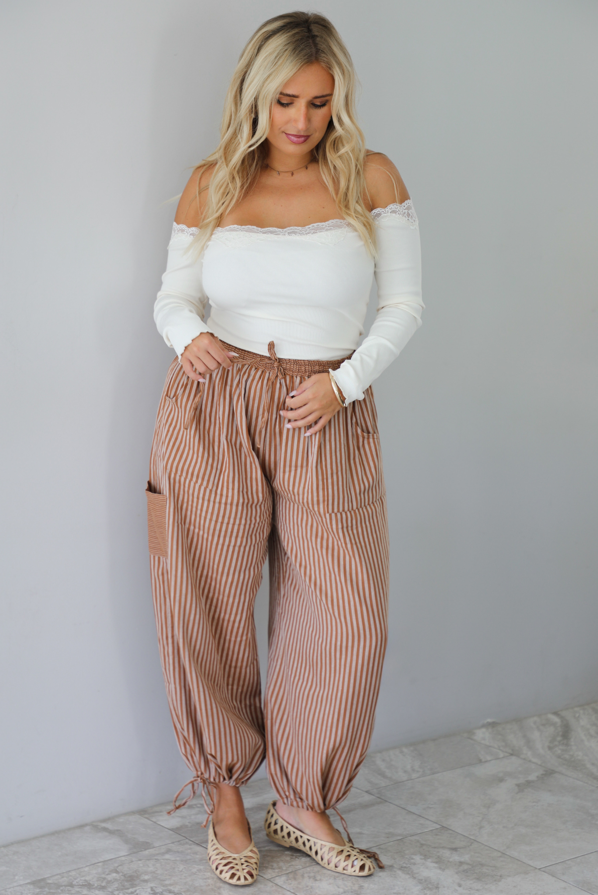 A woman wearing a cream off the shoulder long sleeved top tucked into a pair of camel and cream striped pants, standing indoors against a white wall with white marble flooring. 