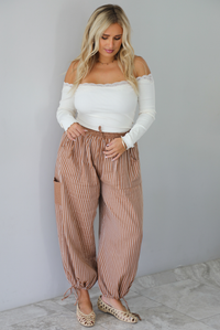 A woman wearing a cream off the shoulder long sleeved top tucked into a pair of camel and cream striped pants, standing indoors against a white wall with white marble flooring. 