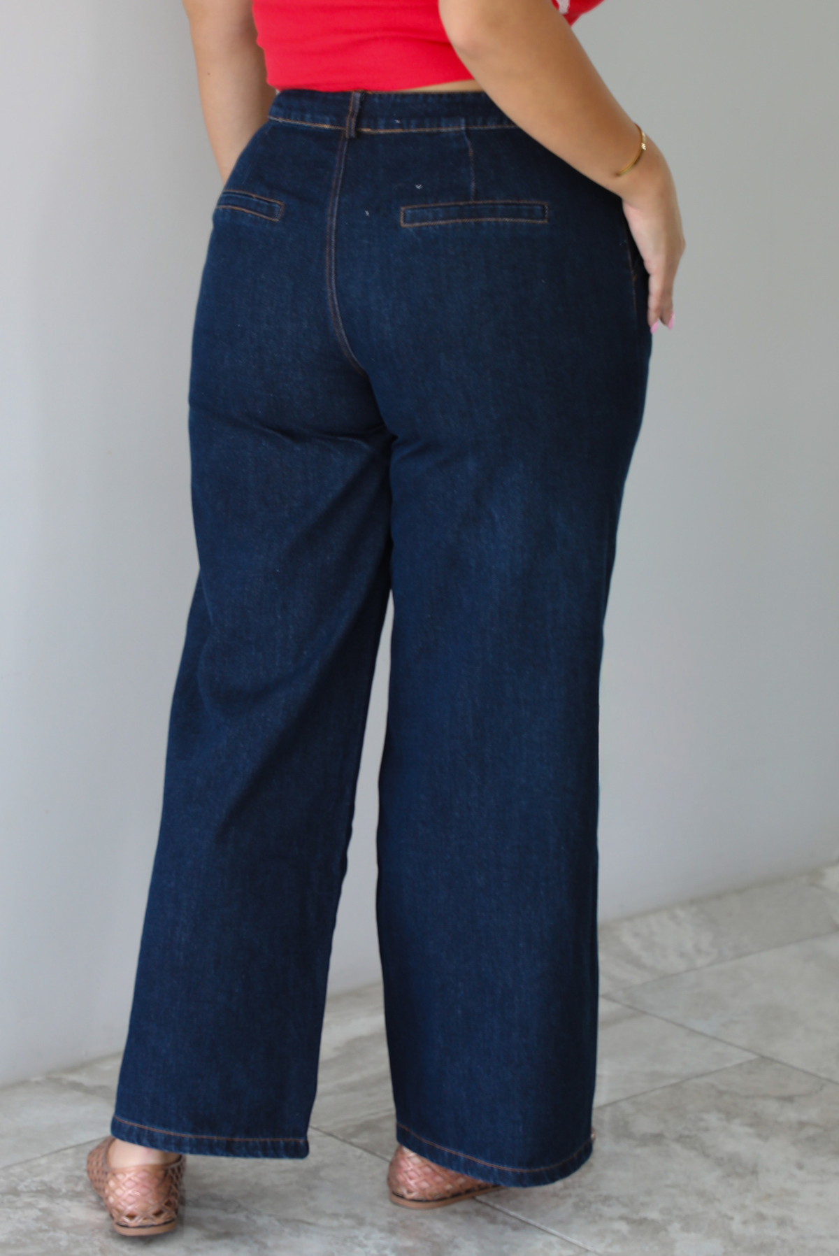 A woman shown from behind wearing wide-leg dark denim jeans, a red top, and brown woven sandals, standing indoors against a white wall with white marble flooring. 